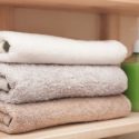 Rolled towels in white, gray, and beige on a wooden shelf, with a green bottle, promoting cleanliness for homeowners.